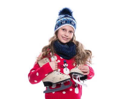Pretty smiling little girl with curly hairstyle wearing knitted sweater, scarf and hat with skates isolated on white background. Winter clothes and sport conceptの写真素材