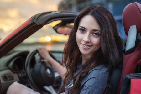 Pretty young brunette woman driving luxury red cabriolet car at the sunset
