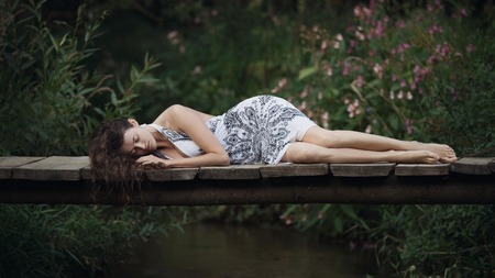 Beautiful young woman lying on the bridge over the forest riverの写真素材