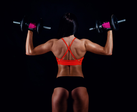 Portrait of an attractive young fitness woman in sportswear doing workout with dumbbells on black background. Tanned sexy athletic girl. A great sporty female body. Back viewの写真素材
