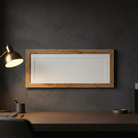 A minimalist home office setup featuring an empty wooden picture frame mounted on a dark gray wall, illuminated by a brass desk lamp. A wooden desk below holds a pen holder and other small items, creating a cozy and productive atmosphere.の素材