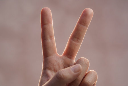 A detailed close-up shot of a human hand displaying the peace sign, with two fingers extended upwards. The background is softly blurred, highlighting the hand and its gesture. The lighting is gentle, creating a subtle glow around the fingers.の素材