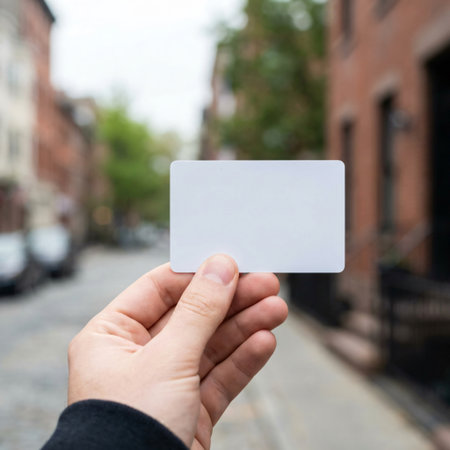 A persons hand extends forward, holding a plain white rectangular card against a blurred background of a city street with buildings and trees, ideal for custom text or branding.の素材