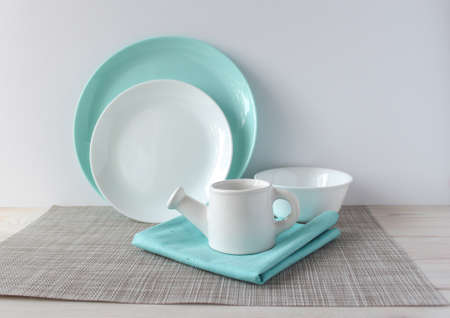 White and turquoise dishes on a woven napkin on a wooden table. Two plates, a bowl, a milk jug in...