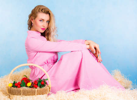 Beautiful young blonde in a long pink dress sitting in a hay over cyan backgroundの写真素材