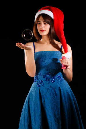 Pretty girl in a blue dress an a Christmas hat catching a soap bubble over black backgroundの写真素材