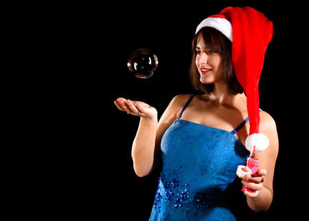 Pretty girl in a blue dress an a Christmas hat catching a soap bubble over black backgroundの写真素材