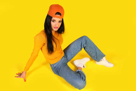 Happy young woman in a casual style clothes sitting on a floor over yellow background
