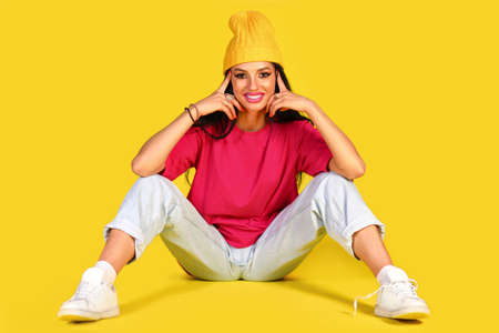 Smiling young woman in a bright pink t-shirt and white jeans sitting on a floor over yellow backgroundの写真素材