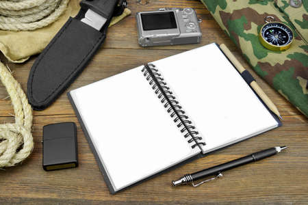 Preparing For Travel. Open Empty Notebook With White Pages, Compact Digital Camera, Rope, Compass, Pen, Pencil, Hunting Knife,  Bag, Cigarette Lighter On Grunge Wood Backgroundの写真素材