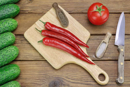 Three Hot Chili Peppers on Wood Cutting Board, One Tomato, Fresh Seven Cucumbers, Salt And Pepper In  The Small Wood Spoon, Kitchen Knife On The Rough Rustic Wooden Tabletopの写真素材