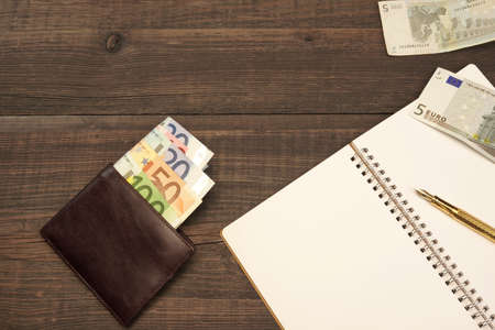 Opened Spiral Notepad With Blank White Page, Golden Fountain Pen And Wallet With Euro Cash On The Rough Wood Table Background With Copy Space, Top Viewの写真素材