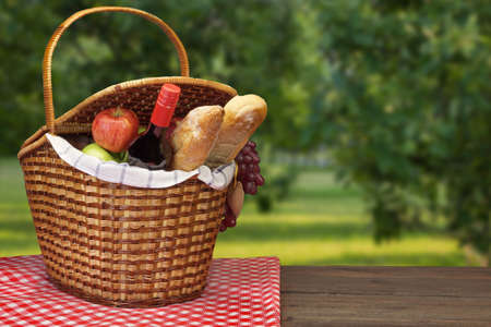Closeup Of Picnic Basket With Food And Wine On The Rustic Brown Table With Red Checkered Tablecloth And  Garden Landscape In The Backgroundの写真素材