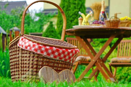 Close-up Of Picnic Basket With Checkered Cloth On The Backyard Lawn. Blurred Outdoor Wooden Table With  Food, Fruits And Champagne Wine Bottle And Yellow House In The Backgroundの写真素材