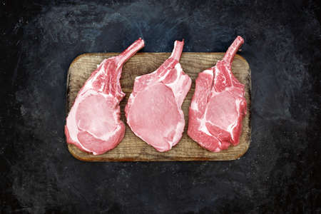 Raw Pork Bone Steaks On Brown Old Cutting Board. Tomahawk Pork Steaks On Chopping Board, Overhead View. Raw Three Pork Steaks On Wood Board And Black Background, Top View.の写真素材