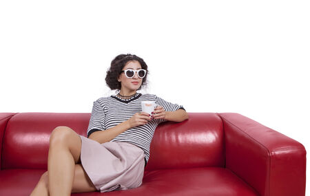 Thoughtful young woman with a coffee cup sitting on a red couch against white backgroundの写真素材