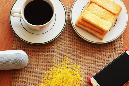 On a brown background there is a black coffee, a crisp bread and a telephone with Bluetooth on itの写真素材