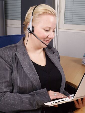 Woman at a helpdesk with white laptop and headset, dressed in a business suit, smiling while watching the PCの写真素材