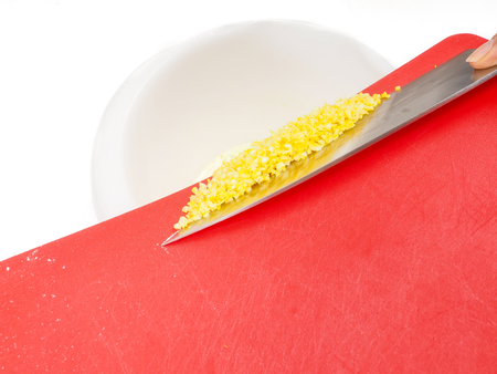 Someone adding chopped lemon zest from a red cutting board with the side of a knife towards whiteの写真素材
