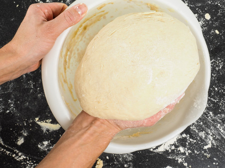 Person lifting a proven dough out from a white plastic bowl on black tableの写真素材