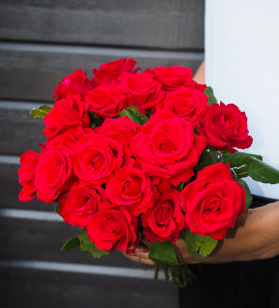 Male person holding a beautiful bouquet of red roses wearing white shirt and black pants against a gray wooden wallの写真素材
