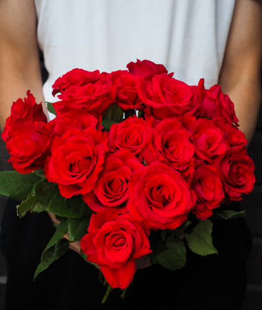Male person holding a beautiful bouquet of red roses wearing white shirt and black pants against a gray wooden wallの写真素材