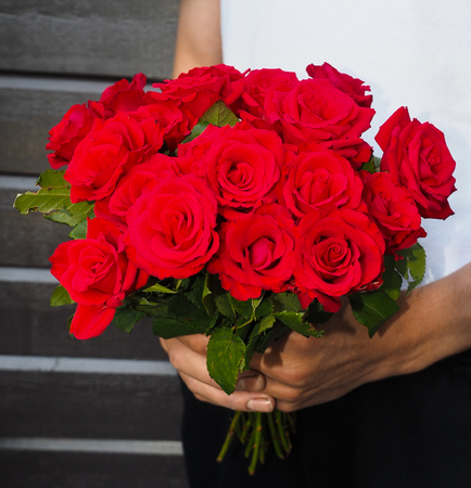 Male person holding a beautiful bouquet of red roses wearing white shirt and black pants against a gray wooden wallの写真素材