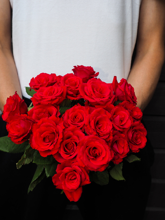 Male person holding a beautiful bouquet of red roses wearing white shirt and black pants against a gray wooden wallの写真素材