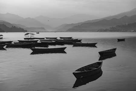 View over a few wooden boats on Phewa Lake in Pokhara, Nepal with the surrounding mountains in the background in black and white. Clear sunny day. Relaxation and calm serenity, peaceful and quiet.の写真素材