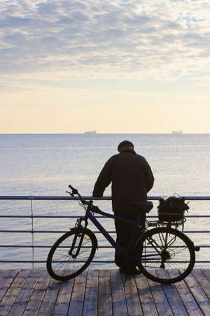 Man with bicycle. Autumn morning seascape with ships on horizon. Silhouette of elderly man on wooden pier. Pastel blue colorsの写真素材