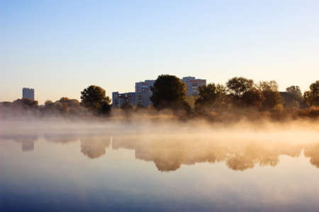 Landscape in city park. Heavy fog on water in morning. Multistory buildings on river bankの写真素材