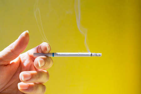 Womans Hand holding a smoldering cigarette against the dark ugly yellow backgroundの写真素材