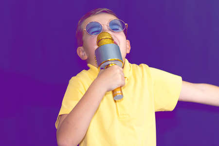 Close up image.The young boy kid child 9s old in yellow t shirt and blue eyeglasses is singing music on purple foggy toned illuminated stage.の写真素材
