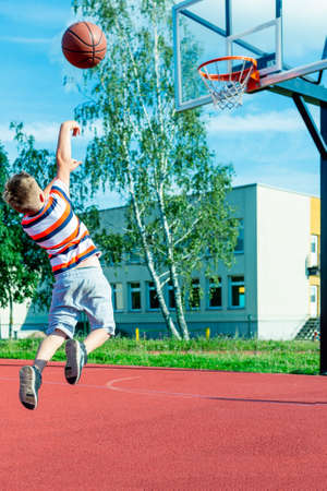 Attractive caucasian little basketball player jumps to throw the ball into the basket hoop on a outdoors red courtの写真素材