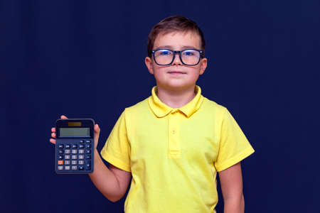 Close up Attractive young schoolboy with eyeglasses, yellow t-shirt holds a calculator.The calculator shows zero. Blue studio background.の写真素材