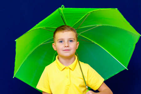 Attractive playful caucasian blond schoolboy with umbrella on blue studio background.Boy shows all kinds of grimaces.close up imageの写真素材