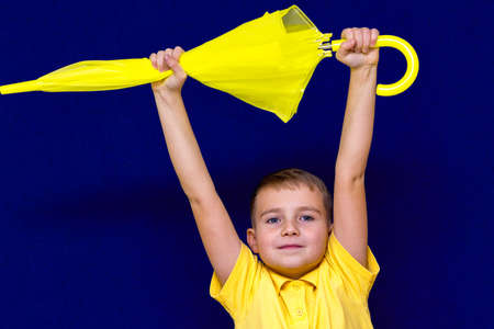 Attractive surprised caucasian blond schoolboy holds unexpanded umbrella on blue studio...