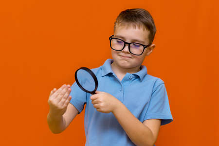 Beautiful european young boy with glasses holding and looking through magnifying glass.Orange studio wall.の写真素材