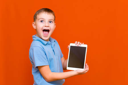 Portrait of surprised schoolboy shows tablet and blue t-shirt on orange studio background.cheerfully thinks something and showing something on empty tablet screen.orange background.Copy space,banner.の写真素材