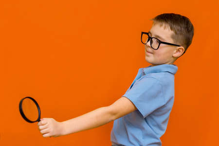 Side view Beautiful european young boy with glasses holding and looking through magnifying glass.Orange studio wall.の写真素材