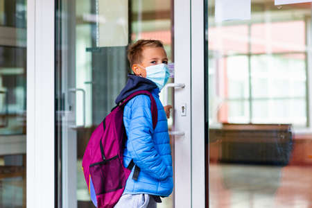 Young schoolboy wearing protective mask is trying to open the school door. Behind the backpack Schoolboy look asideの写真素材