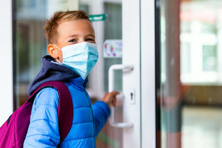 Young schoolboy wearing protective mask is trying to open the school door. Behind the backpack Schoolboy look at cameraの写真素材