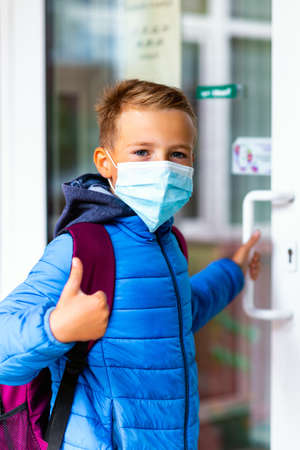 Cute school boy 9s old wears casual clothes and backpack face mask near school. Kid shows thumbs up and holds or opens the door handleの写真素材