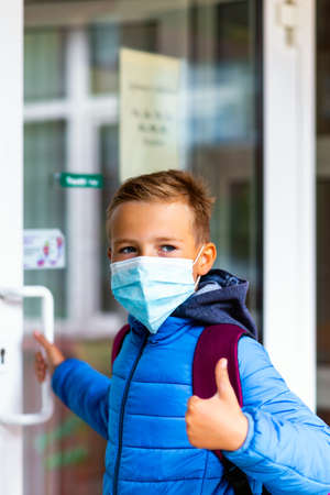 Cute school boy 9s old wears casual clothes and backpack face mask near school. Kid shows thumbs up and holds or opens the door handleの写真素材