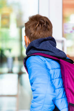 Back view of 9s boy wearing protective mask is trying to open the school door. Behind the backpack Schoolboy looks into the look through the transparent doorの写真素材