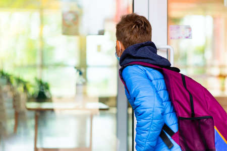 Back view of 9s boy wearing protective mask is trying to open the school door. Behind the backpack Schoolboy looks into the look through the transparent doorの写真素材