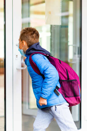 Back view of 9s boy wearing protective mask is trying to open the school door. Behind the backpack Schoolboy looks into the look through the transparent doorの写真素材