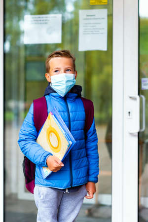Schoolboy wearing protective mask casual clothes and carry a backpack standing near school. Child going school after pandemic over. students are ready for second pandemic waveの写真素材