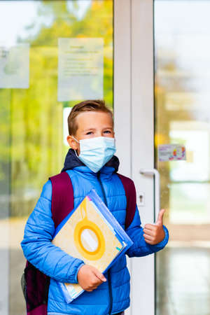 Cute school boy 9s old wears casual clothes and backpack face mask near school. Kid shows thumbs upの写真素材