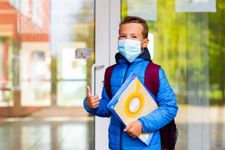 Cute school boy 9s old wears casual clothes and backpack face mask near school. Kid shows thumbs upの写真素材
